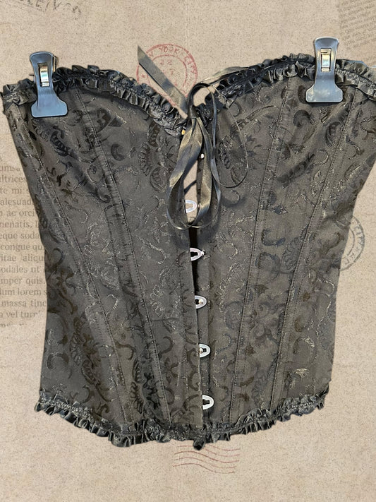 Black floral brocade corset with ruffled trim. Front hook closures and satin bow. Elegant gothic style.