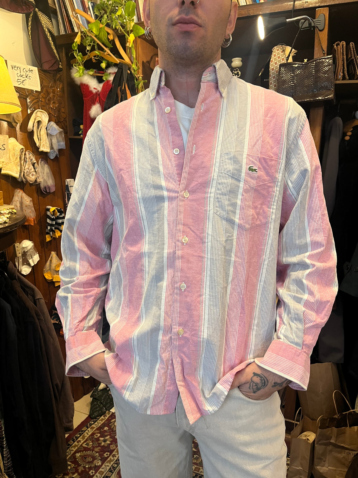 Vintage Lacoste striped button-down shirt in soft pink, grey and white tones, featuring the iconic crocodile logo on the chest pocket. Relaxed fit with a classic button-down collar for an effortless retro look.