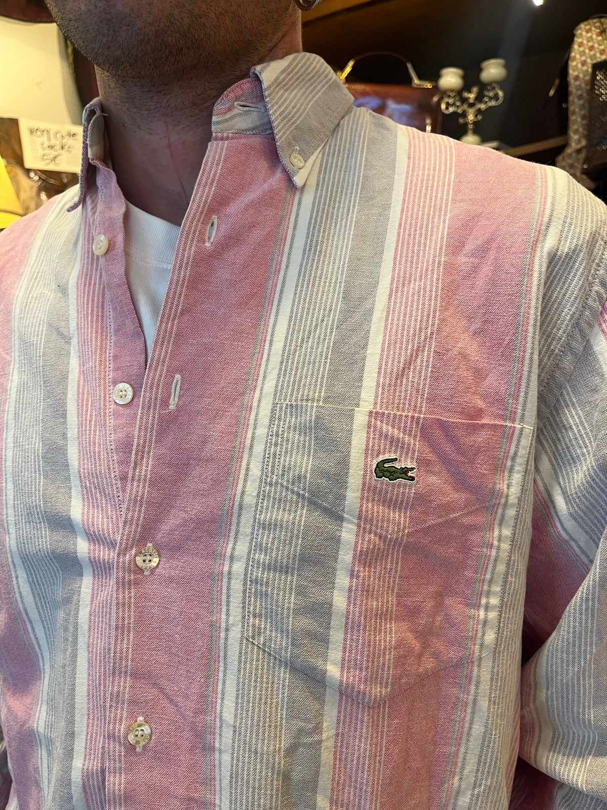 Vintage Lacoste striped button-down shirt in soft pink, grey and white tones, featuring the iconic crocodile logo on the chest pocket. Relaxed fit with a classic button-down collar for an effortless retro look.