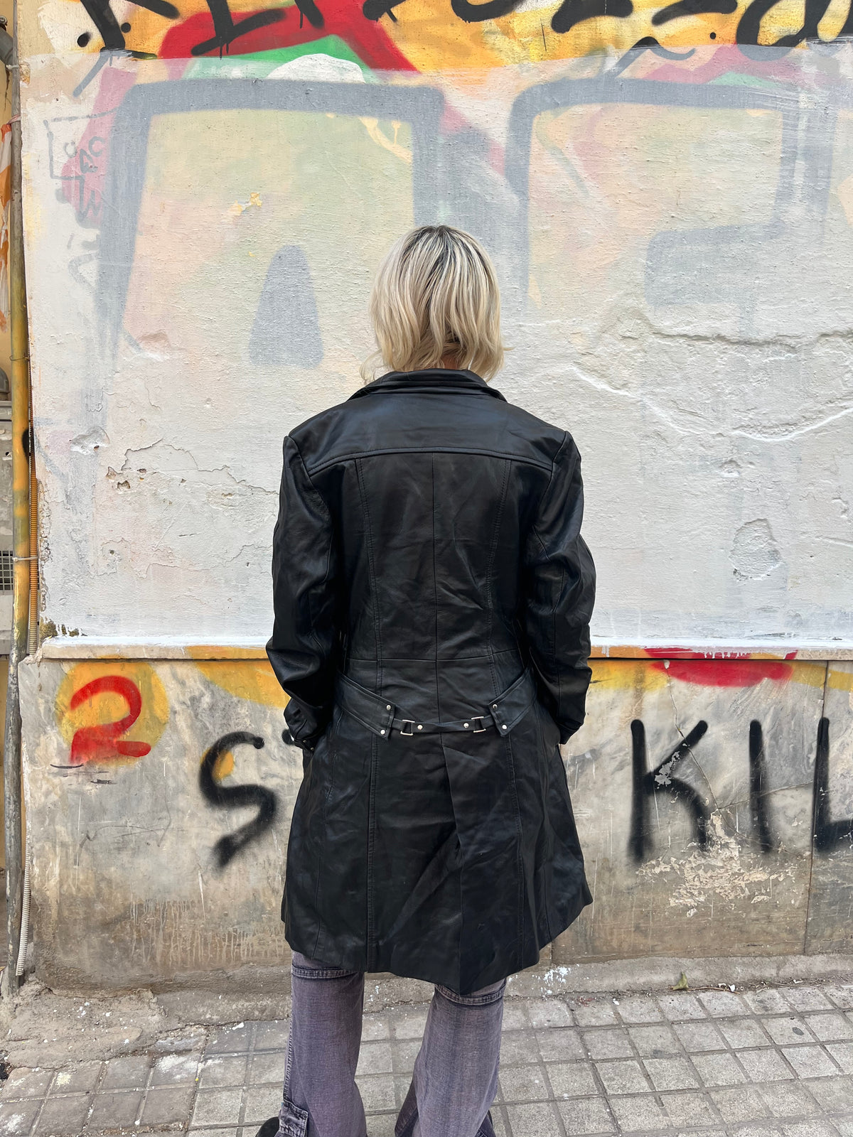 Long black leather coat with a sleek fitted silhouette, full zip closure, and buckle details at the cuffs and back. A bold vintage statement piece with a timeless biker-inspired edge.