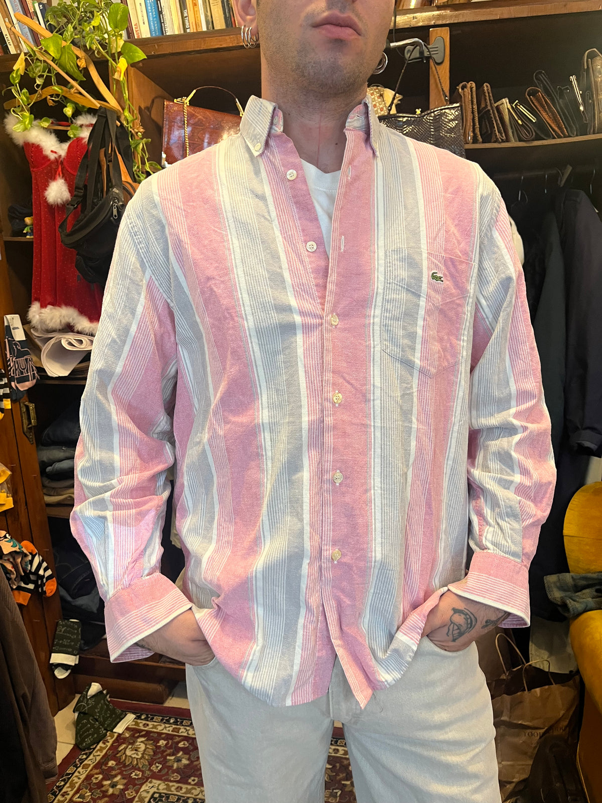 Vintage Lacoste striped button-down shirt in soft pink, grey and white tones, featuring the iconic crocodile logo on the chest pocket. Relaxed fit with a classic button-down collar for an effortless retro look.